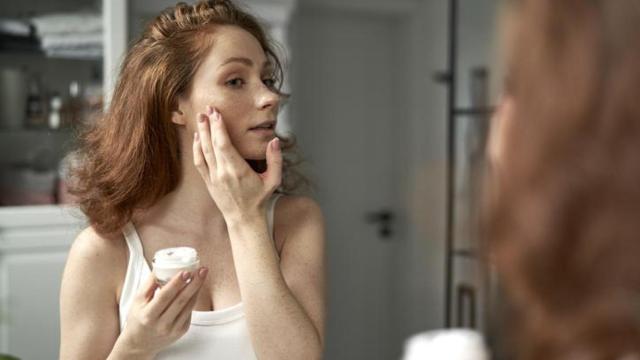 Mujer joven aplicándose crema en el rostro frente al espejo.