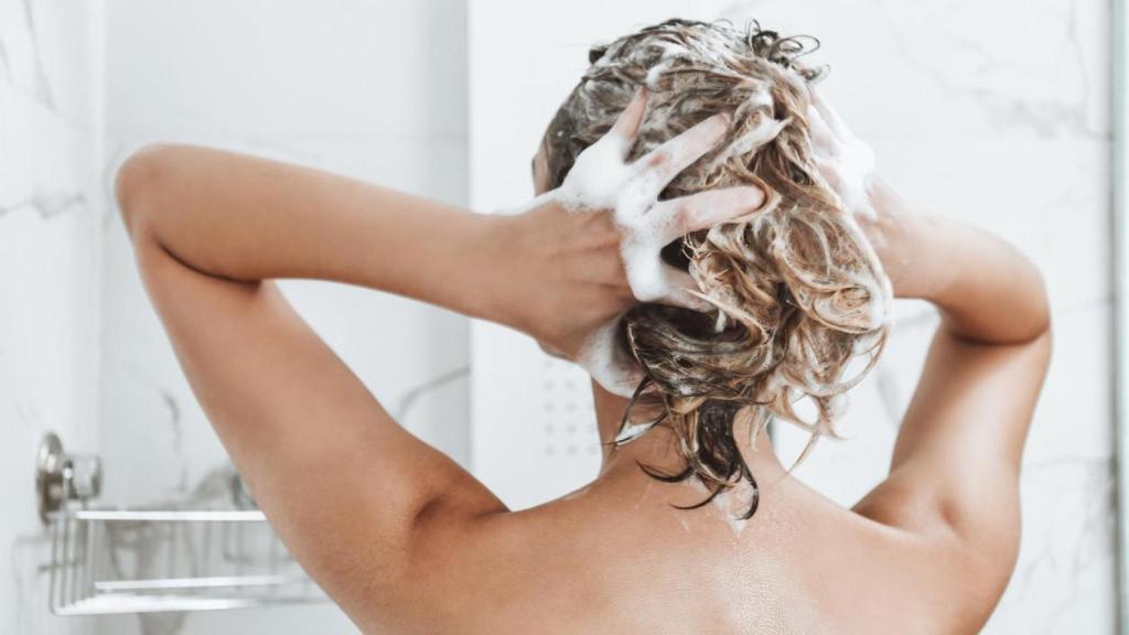 Una mujer de espaldas duchándose y lavándose el pelo.
