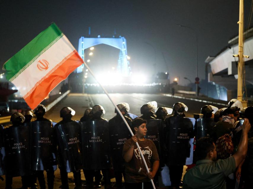 Un niño sostiene una bandera iraní mientras la policía hace guardia durante una protesta contra los ataques israelíes contra Irán en Bagdad.